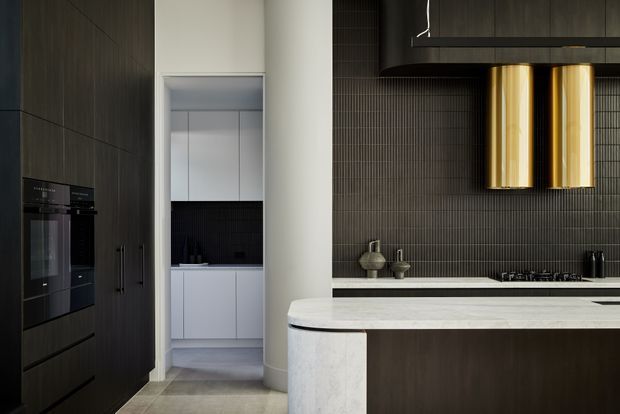 A refined contemporary kitchen with dark vertical-grain cabinetry, integrated black appliances, a large pale stone island with a rounded waterfall edge, and a dark textured tile splashback. Brass cylindrical rangehood elements provide a warm metallic accent. A pantry or secondary preparation area is visible through the opening at the rear.