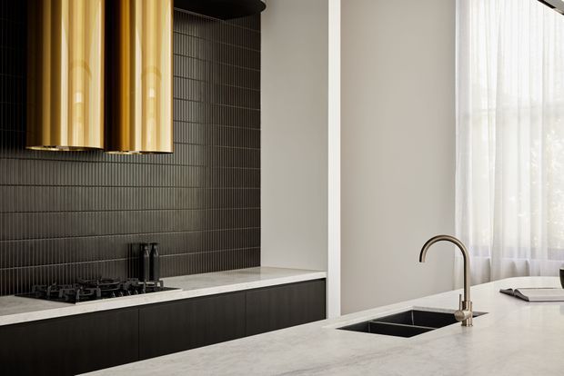 A refined contemporary kitchen featuring a long island with an integrated double-bowl sink and curved brass-toned tap. Dark flat-panel cabinetry and narrow vertical dark wall tiles contrast with a pale stone benchtop and white walls. A large window with sheer curtains provides soft natural light, while a sculptural metallic rangehood forms a strong focal point above the cooktop.