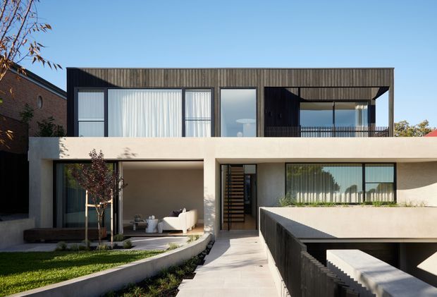 Front exterior view of a contemporary multi-level residence with a strong geometric composition. The house combines a pale rendered lower level with a dark vertically textured upper facade, extensive floor-to-ceiling glazing, deep rectangular overhangs, a recessed balcony, and a landscaped entry path. A partially visible lower-level garage or basement opening appears at the right.