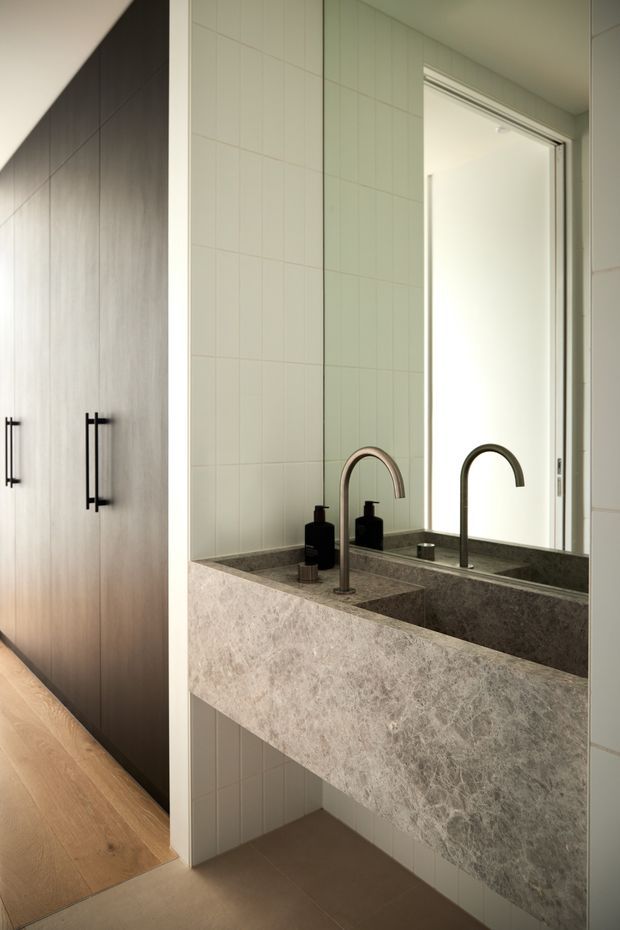 A refined contemporary bathroom vanity viewed beside a dark built-in wardrobe or storage wall. The floating double vanity has a thick grey-veined natural stone basin and benchtop, twin curved black/bronze-toned wall-mounted or deck-mounted faucets, dark soap dispensers, and a large mirror. Pale green-white rectangular wall tiles surround the vanity, with warm timber flooring extending into the adjacent area. The space is softly lit and restrained, combining warm timber, pale tile, dark joinery, and stone.