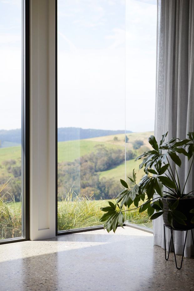 A bright interior corner with floor-to-ceiling glazing overlooking rolling green countryside. The space features a pale terrazzo floor, slender light-coloured window frames, sheer grey curtains, and a large potted plant on a black stand. Natural light creates an airy, calm atmosphere.