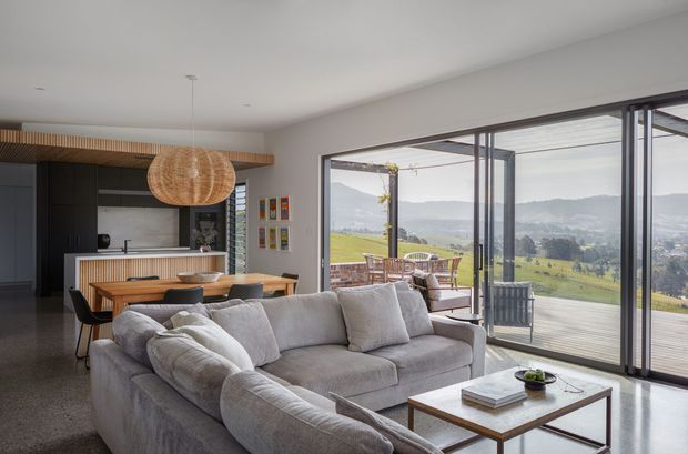 Open-plan contemporary living and dining space with a visible kitchen, featuring a large light-grey sectional sofa, timber coffee table, dining table and chairs, dark kitchen cabinetry, a timber island detail, and a woven pendant light. Full-height glazed sliding doors open to a deck with outdoor dining and expansive rolling green landscape views.