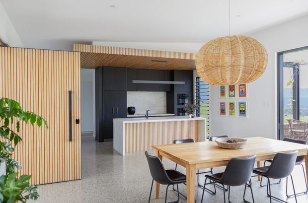 Open-plan contemporary kitchen and dining area with warm timber battens, dark cabinetry, a white island, terrazzo-style flooring, a timber dining table, black dining chairs, and a large woven pendant light. Full-height glazing and a glazed door open toward an outdoor view.