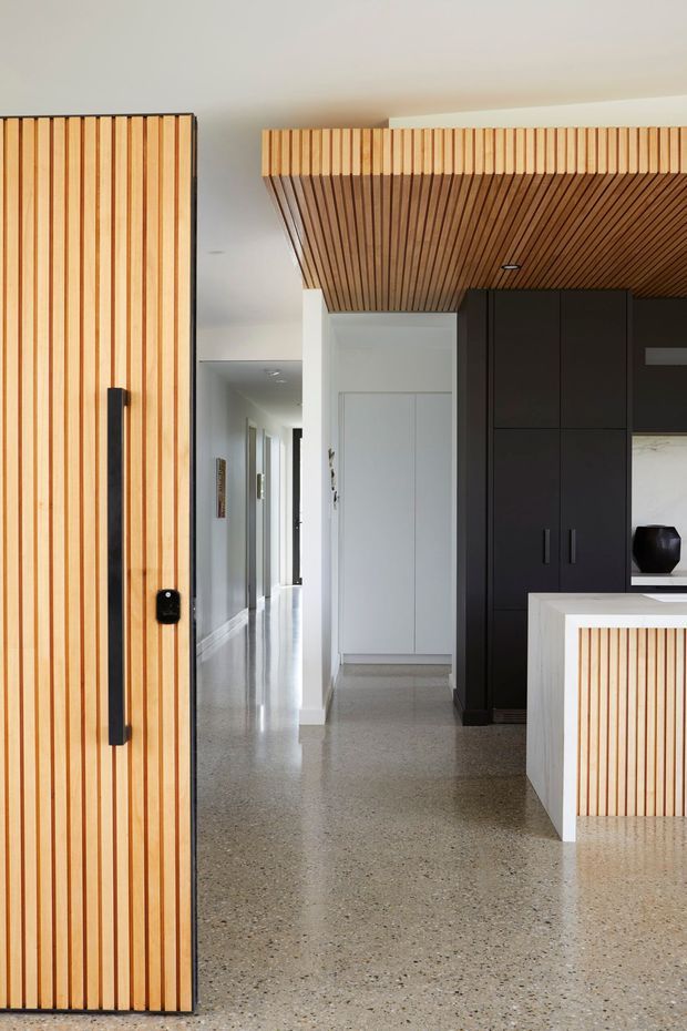 A bright contemporary residential interior showing a kitchen and hallway. The space features extensive vertical and slatted timber detailing on a large pivot door, kitchen island, cabinetry surround, and ceiling, contrasted with matte black cabinetry and crisp white walls. A polished terrazzo or concrete-look floor extends through the open circulation area, with a long hallway visible beyond.