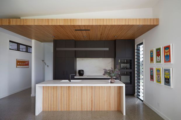 Contemporary kitchen with a large central island featuring vertical timber battens, dark flat-panel cabinetry, a pale stone splashback, and a matching timber-lined ceiling canopy. The space has white walls, polished concrete or terrazzo-style flooring, integrated appliances, louvred windows, and colourful framed artwork.
