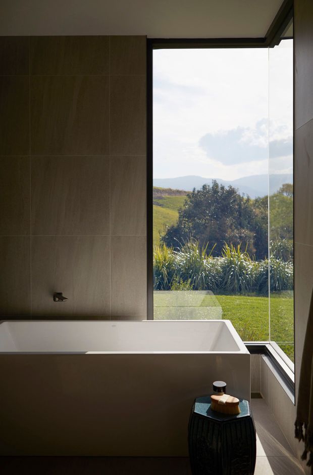 A contemporary bathroom featuring a freestanding white bathtub positioned beside full-height corner glazing. Large grey-brown tiled wall surfaces, dark window frames, and a small dark side table complete the restrained design. The glazing overlooks a landscaped garden and rolling rural hills.