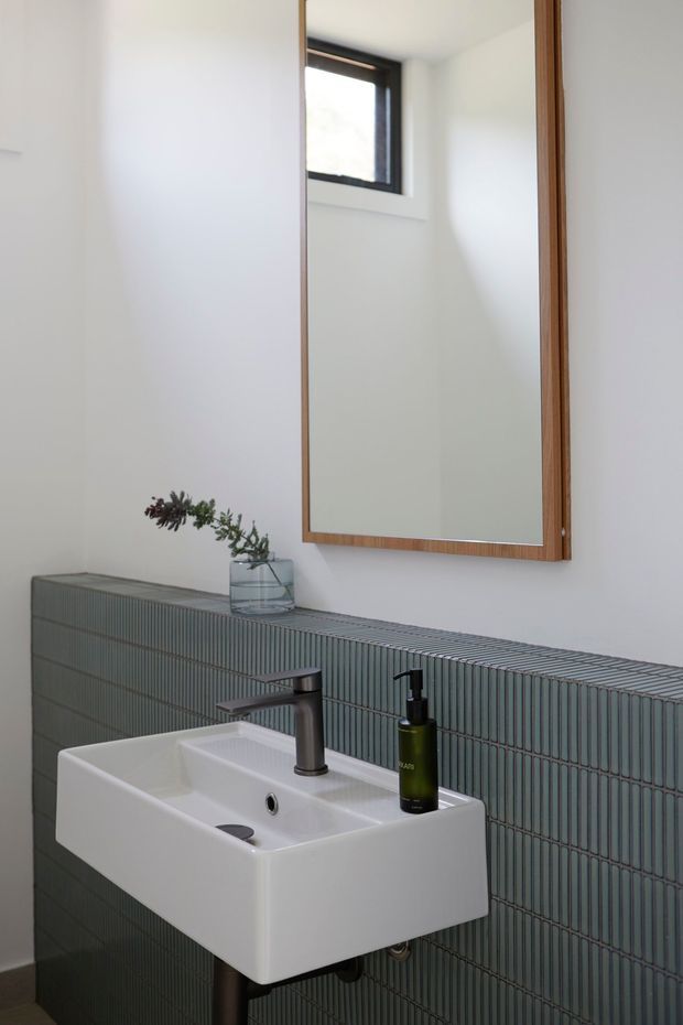 A compact contemporary bathroom featuring a wall-mounted white ceramic basin with a dark metal tap and soap dispenser. The lower walls are clad in vertically ribbed blue-grey tiles, while the upper walls are white. A large timber-framed mirror, small high-set window, and simple glass vase complete the understated design.