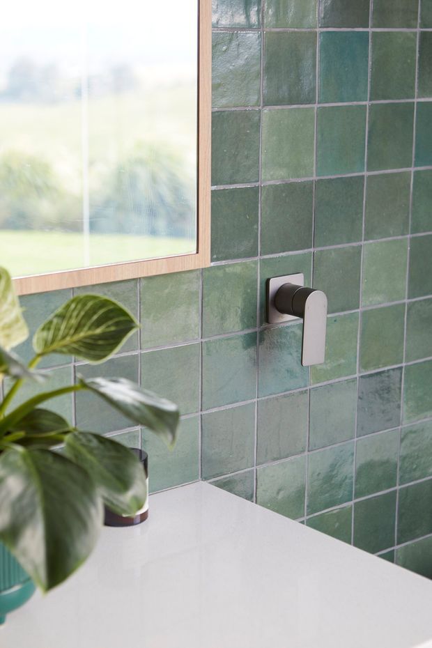 Close-up of a refined bathroom vanity area with a glossy white benchtop, green square tiled wall, wall-mounted minimalist tap, timber-framed mirror, and leafy indoor plant. Soft natural light enters through the mirror-side window, creating a calm, organic atmosphere.