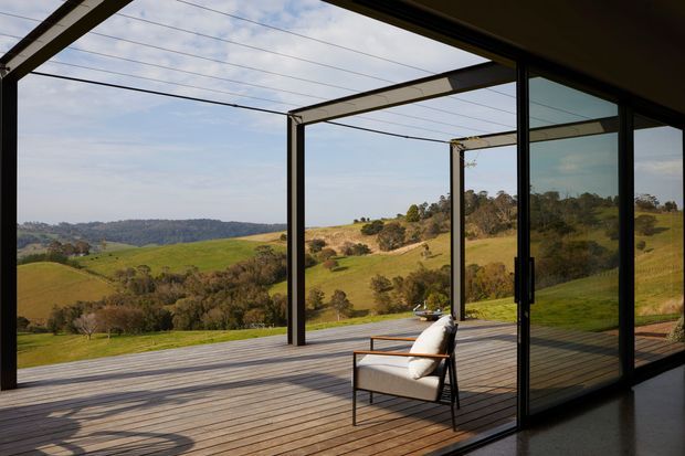 A minimalist indoor-outdoor living space with full-height sliding glass doors opening onto a spacious timber deck. Slim dark metal framing and exposed structural beams create a strong connection to the surrounding rolling green rural landscape, with a single modern outdoor lounge chair on the deck.