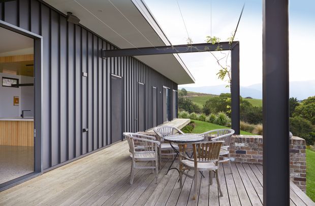 A contemporary outdoor deck and terrace beside a dark vertically clad building. The timber decking is furnished with a round dining table and chairs beneath a simple overhead frame, with a partially visible interior through glazed doors on the left. A low brick retaining wall and landscaped rural or coastal views extend beyond the deck.