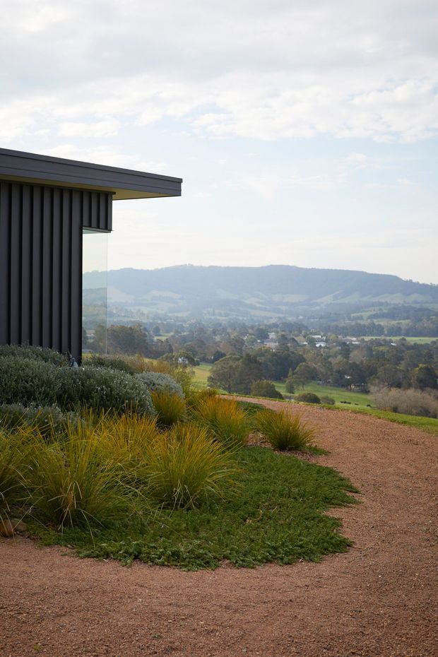A contemporary dark-clad building edge with a projecting flat roof and full-height glazing overlooks a broad rolling rural landscape. Native-style grasses, low planting, and a reddish gravel path occupy the foreground, creating a strong connection between the minimal architecture and the surrounding countryside.