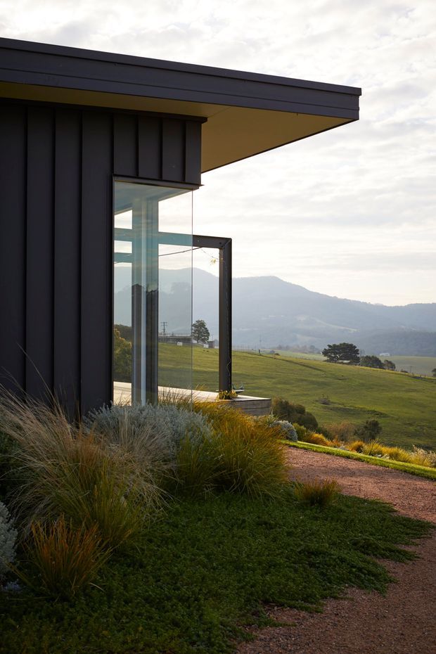 A contemporary single-storey pavilion-like building set on a sloping rural landscape. The dark vertically clad facade has a broad flat projecting roof and expansive frameless corner glazing, reflecting the surrounding hills. Native-style grasses and low groundcover surround a gravel path, with distant mountains visible beyond.
