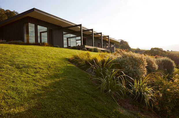 A low, elongated contemporary residence set into a sloping landscaped site. The building features dark vertical cladding, broad floor-to-ceiling glazing, and a projecting roof with a repeating structural frame along the terrace. A manicured lawn and layered native-style planting surround the house, with warm low-angle sunlight and an expansive distant outlook.