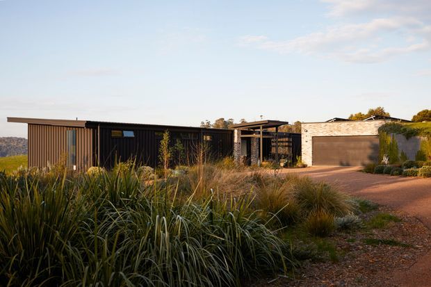 A low, single-storey contemporary residence viewed from the landscaped front approach. The home combines dark vertical cladding and extensive glazing with a projecting flat roof and a separate pale masonry garage volume. Native grasses and drought-tolerant planting frame a gravel and paved driveway, creating a restrained, landscape-integrated exterior.
