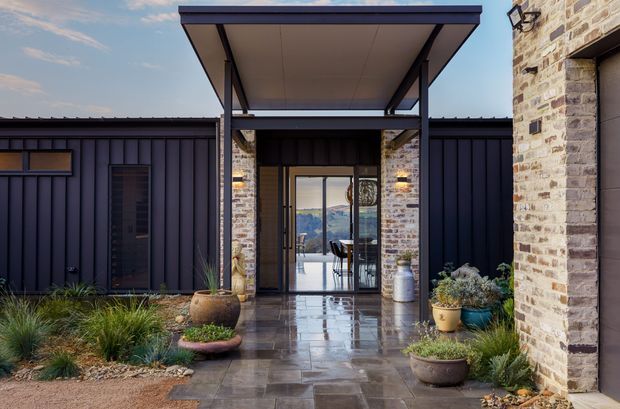 Exterior view of a contemporary residential entrance and covered walkway. Dark vertical metal cladding frames a glazed entry, with a large flat-roofed canopy supported by slender dark steel posts. Pale rough stone masonry forms the side wall and entry surround. The paved approach includes gravel, drought-tolerant ornamental planting, and ceramic planters. Through the glazing, a dining area and distant landscape are visible.