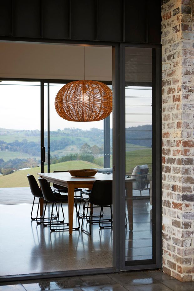 A refined dining area viewed through dark-framed glazed doors, with a rectangular timber dining table, black chairs, and a large woven rattan pendant light. The room features polished concrete flooring, exposed brickwork, dark metal wall cladding, and expansive glazing overlooking rolling green countryside. A timber deck is visible beyond the dining space.