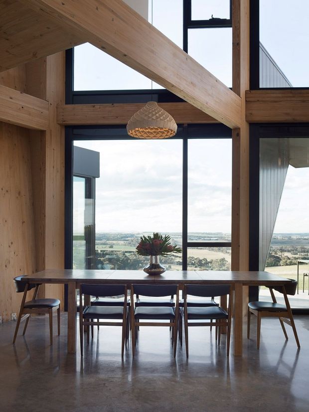 A double-height dining area with a long timber dining table and mid-century style chairs, framed by expansive black-framed glazing overlooking a rural landscape. Exposed timber post-and-beam structure creates a strong diagonal ceiling feature, with a woven pendant light above the table and polished concrete flooring below.