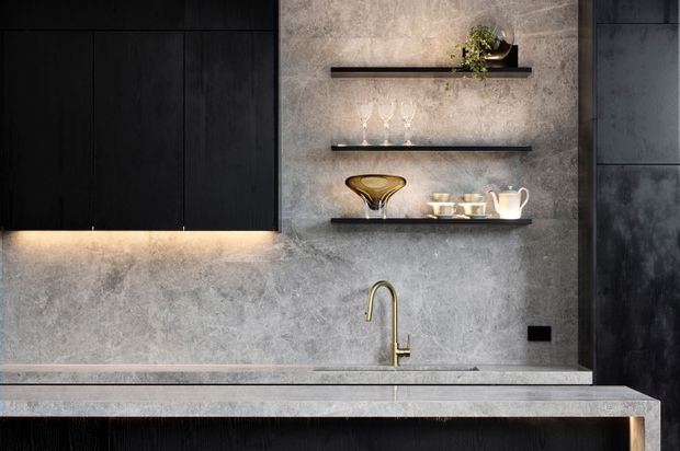 A close-up view of a sophisticated contemporary kitchen or bar joinery area. Dark matte cabinetry occupies the left side, while a grey textured stone-look wall and matching benchtop frame three slim open shelves displaying glassware and ceramics. A brass-toned curved tap and undermount sink sit in the benchtop, with concealed warm LED lighting beneath the cabinetry and shelves. The composition is highly refined, dark, and minimalist with strong horizontal lines.