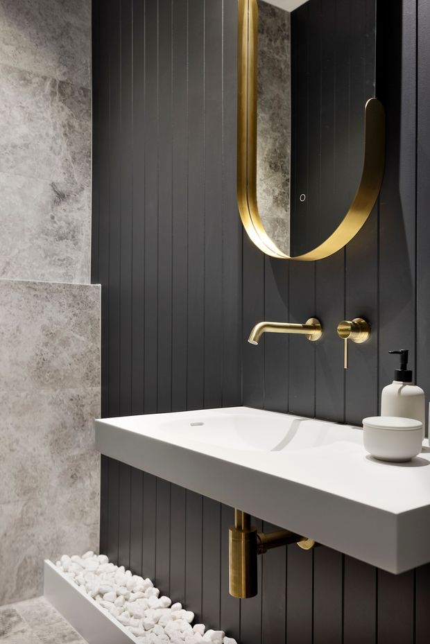A compact, high-contrast bathroom vanity featuring a floating white rectangular basin, matte black vertically grooved wall panelling, pale grey stone-look wall and floor tiles, and brushed brass/gold tapware, exposed drain, and circular mirror frame. White decorative pebbles sit in a narrow built-in floor trough beneath the vanity, adding a sculptural natural detail.