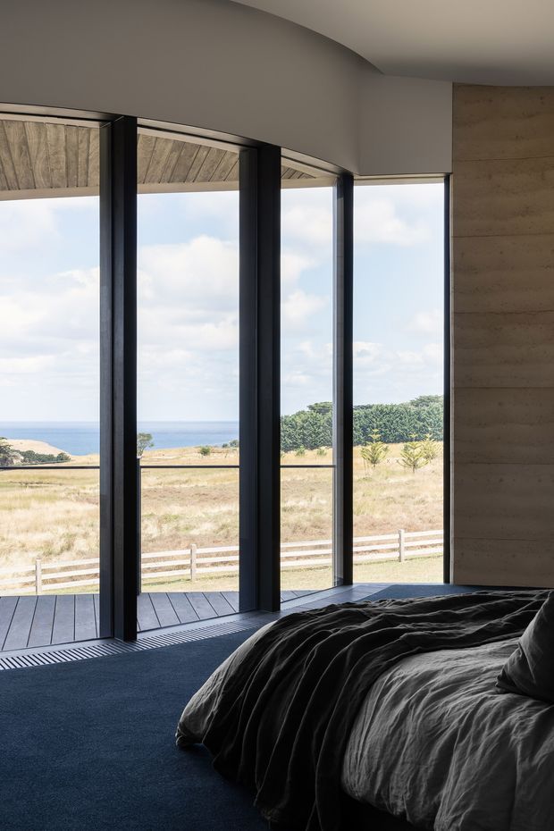 A contemporary bedroom photographed from beside the bed, with a dark upholstered bed and rumpled charcoal bedding in the foreground. Floor-to-ceiling black-framed glazing opens to a deck and a broad coastal landscape with ocean views. The room has a curved smooth plaster ceiling and a textured concrete or rammed-earth feature wall, creating a restrained, nature-connected atmosphere.