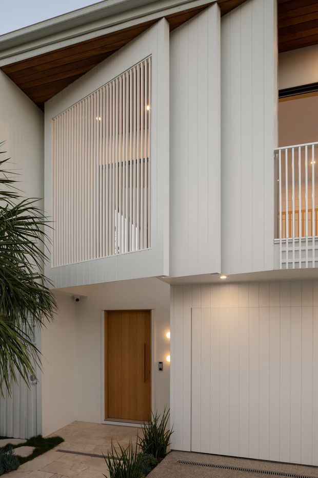A close-up exterior view of a contemporary multi-level home entrance and facade. The building features white vertical panel cladding, a warm timber-lined sloping soffit, a timber front door, and tall white vertical screening over upper-level glazing and balcony areas. Small landscaped planting and pale stone paving frame the entry.