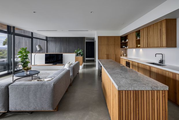 Open-plan contemporary kitchen and living area with a large timber-slatted island and matching upper cabinetry, grey stone benchtops, dark timber wall panelling, polished concrete flooring, and full-height black-framed glazing. A grey sectional sofa and media console occupy the living zone, with greenery and a landscape visible beyond the windows.
