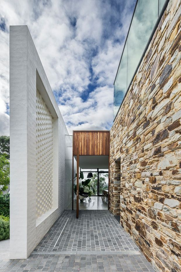 A narrow modern architectural entry passage framed by a crisp white rendered wall on the left and a rugged natural stone wall on the right. A timber-clad overhead entry volume leads toward glazed interiors with visible planting and furniture. The passage is paved in small grey modular tiles, while the composition combines strong rectilinear forms, textured masonry, glass, and timber beneath a partly cloudy sky.