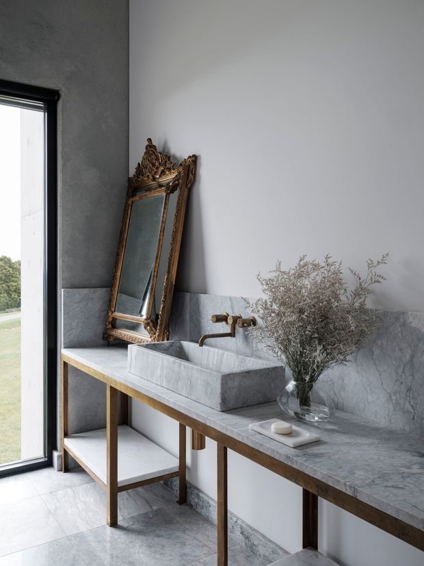 A refined bathroom vanity area with a large rectangular stone basin set into an open-frame console, pale grey marble or stone wall and benchtop surfaces, and a wall-mounted brass tap. An ornate antique gold-framed mirror leans against the wall beside a glass vase with dried foliage. The space is softly lit by a tall black-framed window and has a restrained, luxurious contemporary-classic character.