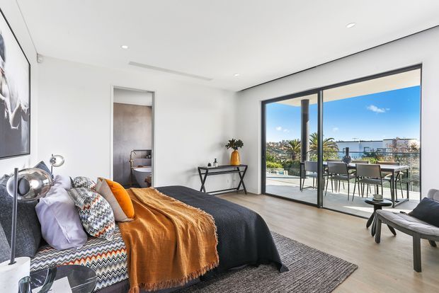 Bright contemporary bedroom with a neatly made bed, layered patterned and neutral cushions, an ochre throw, slim console table, and minimalist decor. Large black-framed sliding glass doors open to a furnished balcony or terrace with an urban coastal outlook. An ensuite bathroom is visible through the open doorway.