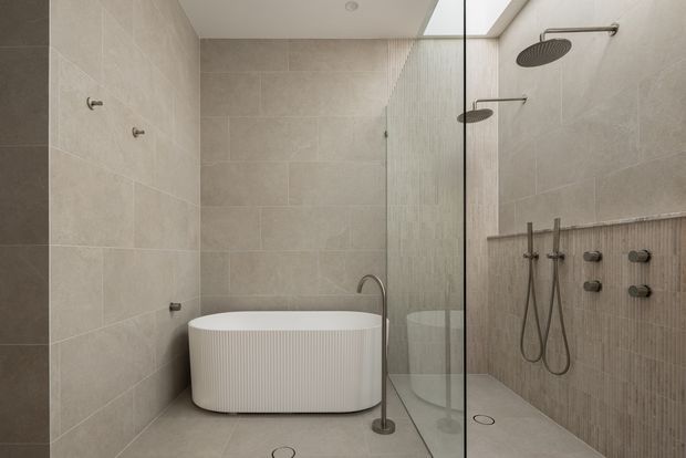 Contemporary bathroom featuring a freestanding white ribbed bathtub, large warm grey stone-look wall and floor tiles, a frameless glass shower enclosure with dual rainfall showerheads and handheld controls, and a narrow skylight providing soft natural light. A vertically textured tile feature wall adds subtle detail.