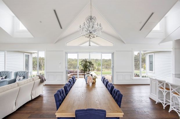 Bright, spacious open-plan dining and living area with a long timber dining table surrounded by navy upholstered chairs, white paneled walls, large windows, and a dramatic vaulted ceiling featuring a crystal chandelier. A lounge seating area is visible to the left and a white built-in bar or kitchen joinery area is visible to the right.