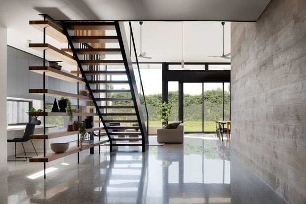 A bright, open-plan contemporary interior featuring a sculptural open-riser staircase with dark metal stringers and timber treads, integrated open shelving, polished concrete flooring, a textured concrete feature wall, and expansive black-framed glazing overlooking greenery. Minimal furnishings and abundant natural light create a refined, airy atmosphere.