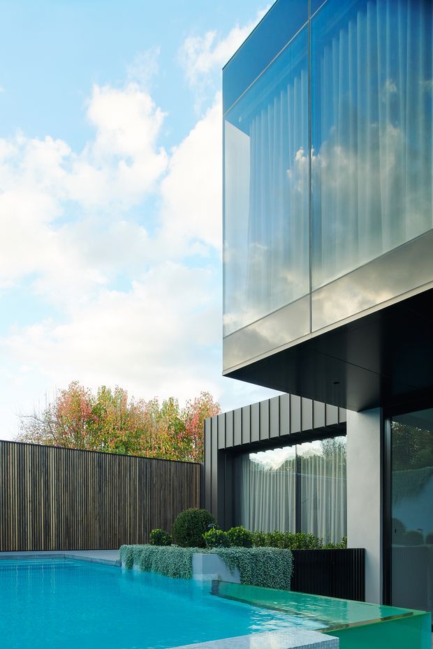 Contemporary two-storey residential exterior viewed from a private pool courtyard. The building features a projecting upper volume clad in reflective blue-grey glazing, dark soffits and walls, vertical metal cladding around the lower level, and large glazed openings. A turquoise swimming pool, raised planted beds, clipped shrubs, and a tall timber privacy fence define the landscaped outdoor setting.