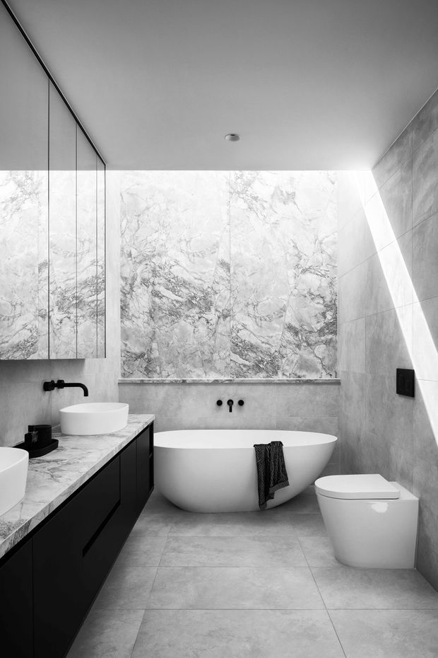 A refined contemporary bathroom in a monochrome palette. Large grey porcelain-look floor and wall tiles frame a freestanding white oval bathtub, wall-mounted white toilet, and black vanity cabinetry with a pale veined stone benchtop and vessel basins. A large veined marble-look feature wall sits behind the bath, while a full-height mirror and slim black fixtures add contrast. Natural light enters from the right, creating a bright diagonal highlight.
