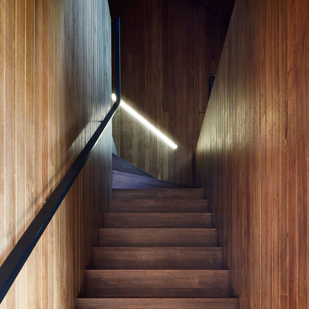 A narrow, atmospheric stairway enclosed by rich vertical timber wall panelling. Dark timber stair treads rise toward a landing or turn, with a slim black handrail along the left wall and a linear integrated light creating a bright diagonal accent. The composition is minimalist and highly crafted, with warm wood dominating the enclosed space.