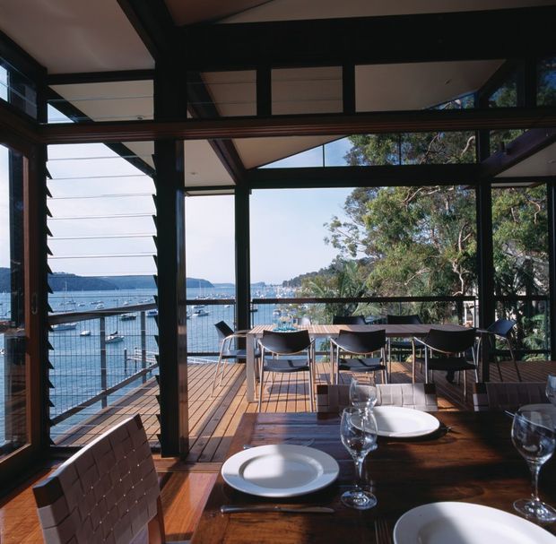 A glazed dining area opening onto a timber deck, with a long dark timber dining table set with white plates and wine glasses. Black-framed glazing and louvred windows create an airy indoor-outdoor connection, with a waterfront marina and tree-covered shoreline visible beyond.