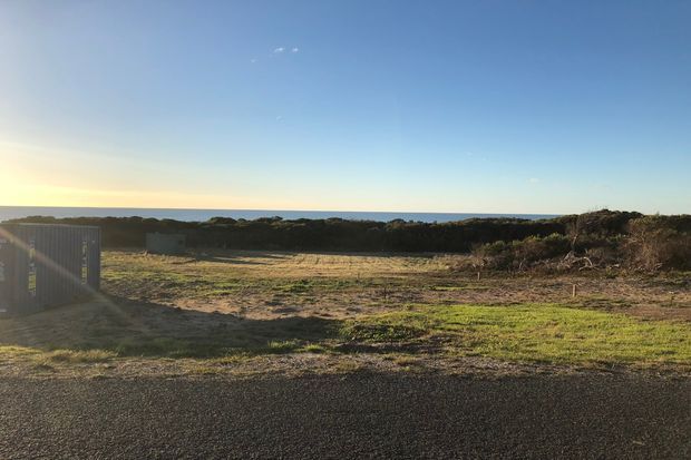 A wide outdoor coastal landscape viewed from a paved area, with low scrub vegetation, sandy open ground, a distant ocean horizon, and clear blue sky. A dark slatted structure or fence is partially visible at the left edge. The image is a straightforward exterior site view with warm low-angle sunlight.