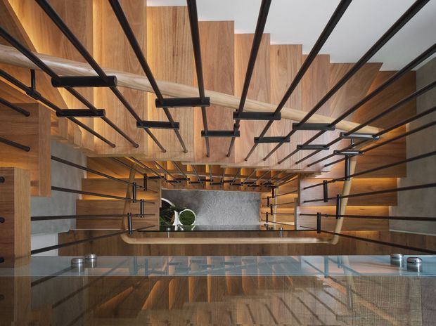 A dramatic interior view looking across a glass balustrade into a multi-level stairwell or landing. Warm timber wall and ceiling panelling forms strong geometric planes, contrasted by extensive black metal rod railings and structural elements. A lower-level area with a small seating or decorative arrangement is visible below, along with reflective glass surfaces. The composition emphasizes vertical circulation, layered platforms, and bold linear detailing.