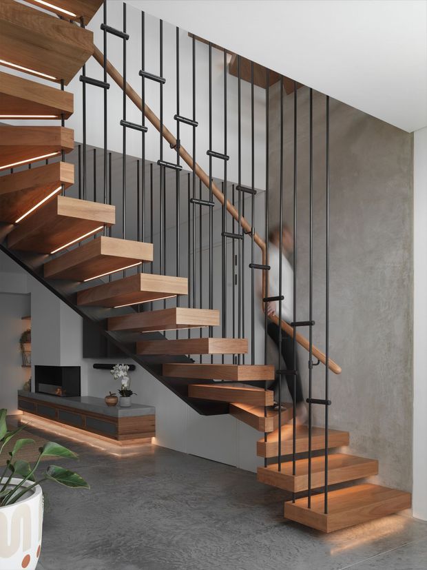 Interior view of a contemporary open-plan living area dominated by a sculptural floating staircase. Thick timber stair treads with integrated warm LED lighting are supported by a dark structural spine and enclosed by slender black vertical metal balusters, with a timber handrail. The staircase rises alongside a textured grey concrete-look wall and a dark grey feature wall. A low timber media console and television sit beneath the stairs, while a potted plant occupies the foreground. The space has a refined, minimalist material palette of timber, dark metal, concrete-grey surfaces, and white ceilings.