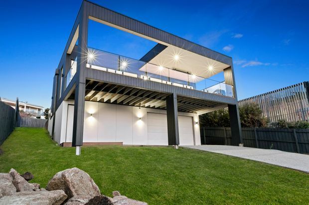 Exterior view of a contemporary multi-level residence with a bold dark framed structure, open upper-level terrace, extensive glazing, covered ground-floor parking or outdoor space, and a flat roof. The home sits on a sloping landscaped site with lawn, boundary fencing, and exposed rock landscaping.