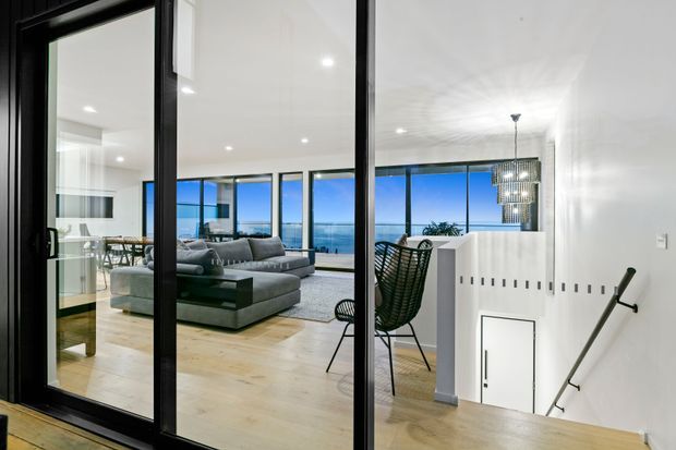 View through black-framed glass doors into a bright contemporary open-plan living and dining area. The space features pale timber flooring, white walls and cabinetry, a large grey sectional sofa, dining furniture, a sculptural pendant light, and expansive glazing overlooking the ocean. A stairwell with a simple black handrail is visible in the foreground.