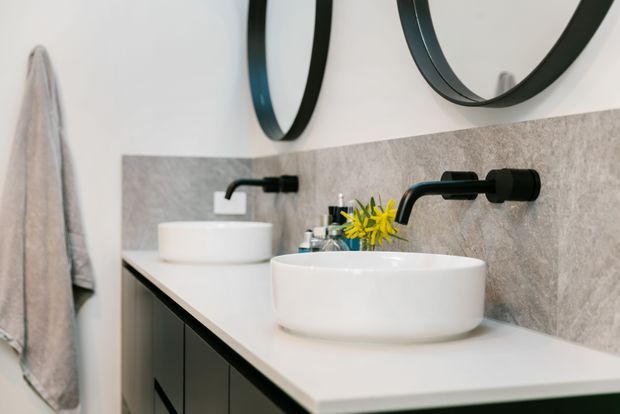 Contemporary double-basin bathroom vanity with two white vessel sinks, matte black wall-mounted tapware, dark cabinetry, a pale benchtop, and a grey stone-look splashback. Round black-framed mirrors and a hanging towel are visible.