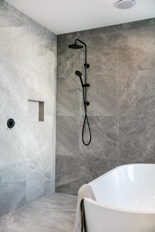 Contemporary bathroom with a walk-in shower clad in large-format grey stone-look tiles, matte black rain shower and handheld fixture, recessed wall niche, and a partially visible freestanding white bathtub. The room has a clean, refined, spa-like appearance with a white ceiling and discreet ceiling vent.