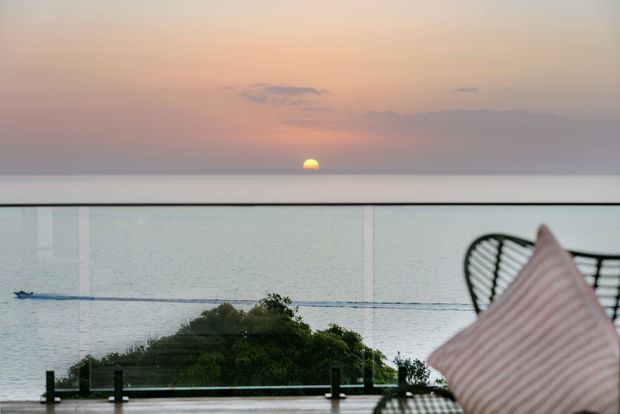 A tranquil sunset or sunrise ocean view seen from a modern balcony or terrace. The foreground features a frameless glass balustrade with dark posts, a leafy tree below, and a black wire outdoor chair with a pale striped cushion at right. The muted pink, peach, grey, and blue horizon creates a calm coastal atmosphere.