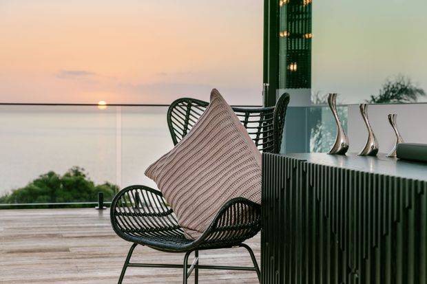 A refined outdoor terrace or balcony at sunset, featuring a black woven lounge chair with a textured blush cushion, a matching chair behind it, and a dark vertically fluted sideboard topped with sculptural metallic decor. Full-height glazing and a transparent balustrade overlook water and greenery, creating a calm, elevated setting.