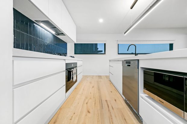 Bright contemporary kitchen with white handleless cabinetry, a pale timber floor, dark blue-green glazed tile splashback, integrated stainless-steel appliances, and a central island with sink. Narrow horizontal windows and linear ceiling lighting contribute to a clean, minimalist look.