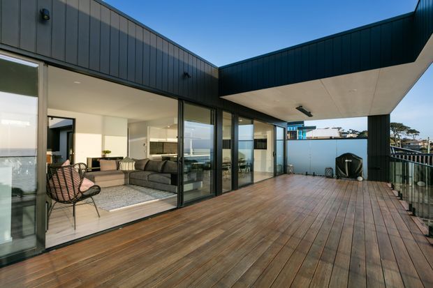 A contemporary rooftop-style outdoor deck adjoining a glass-fronted living area. The home has dark vertical metal cladding, expansive sliding glazing, a broad timber deck, and a projecting flat roof canopy. Through the glazing, a living room and kitchen are visible, with a coastal outlook beyond.