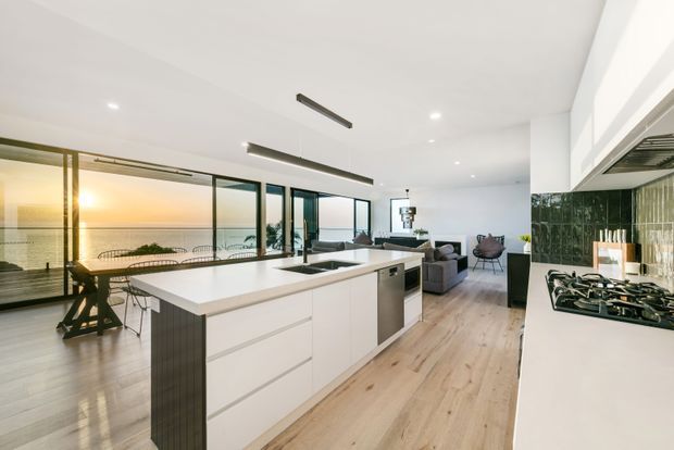 Open-plan contemporary kitchen and living area with a large white island, integrated appliances, dark green tiled splashback, light timber flooring, and expansive glazing overlooking the water. A dining area and living room extend beyond the kitchen, with linear pendant lighting and a bright, refined coastal outlook.