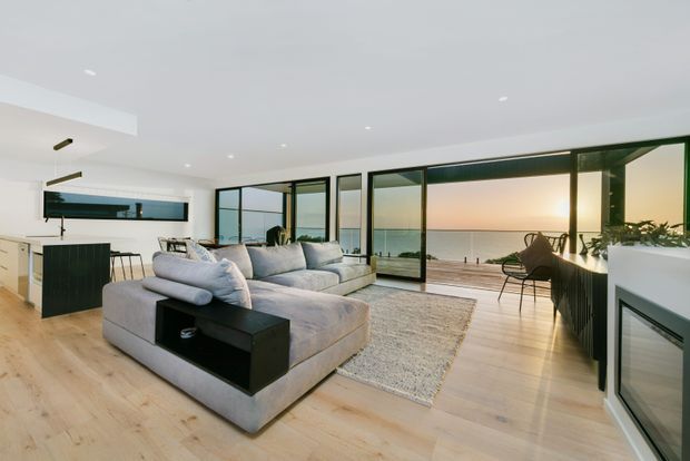 Bright open-plan living room with a large light-grey sectional sofa, black side table, pale timber flooring, and expansive black-framed glazing opening to a waterfront terrace. A compact kitchen with a dark island is visible to the left, while a fireplace or built-in feature is visible on the right. The room has a clean, contemporary coastal outlook with sunset views.