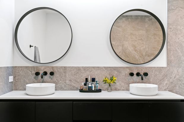 Contemporary double-vanity bathroom with two round black-framed mirrors, white vessel basins, matte black wall-mounted tapware, a pale stone-look tiled splashback and a long dark vanity with integrated storage. Small toiletries and flowers sit on the benchtop.